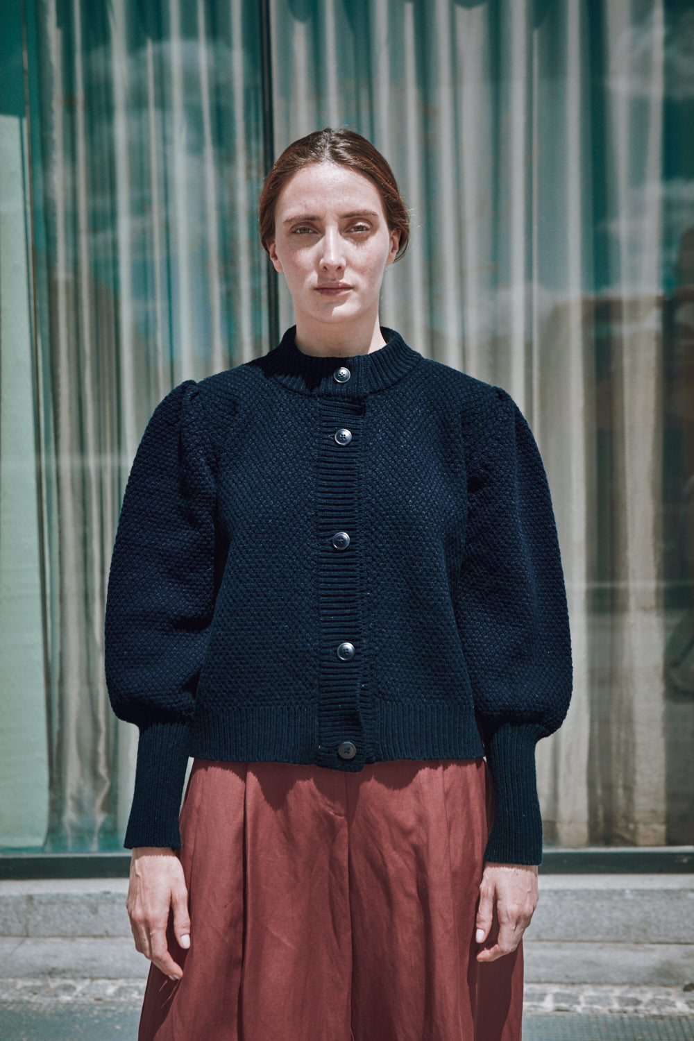 Woman wearing a dark blue L'ENVERS cardigan and red skirt standing in front of a glass building.